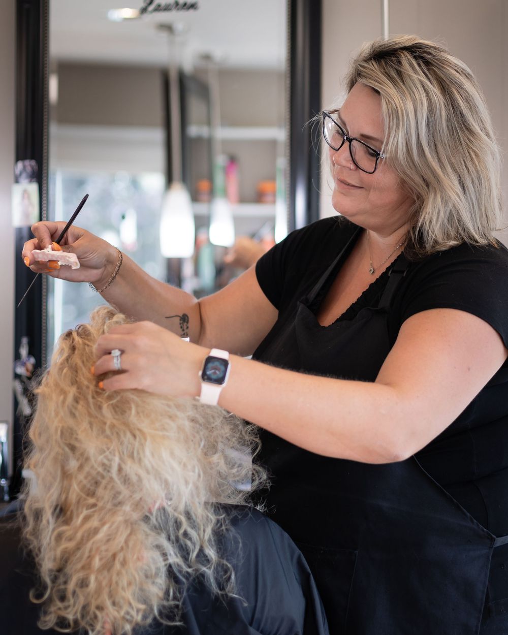 Hair stylist working on client's curly blonde hair in professional salon.