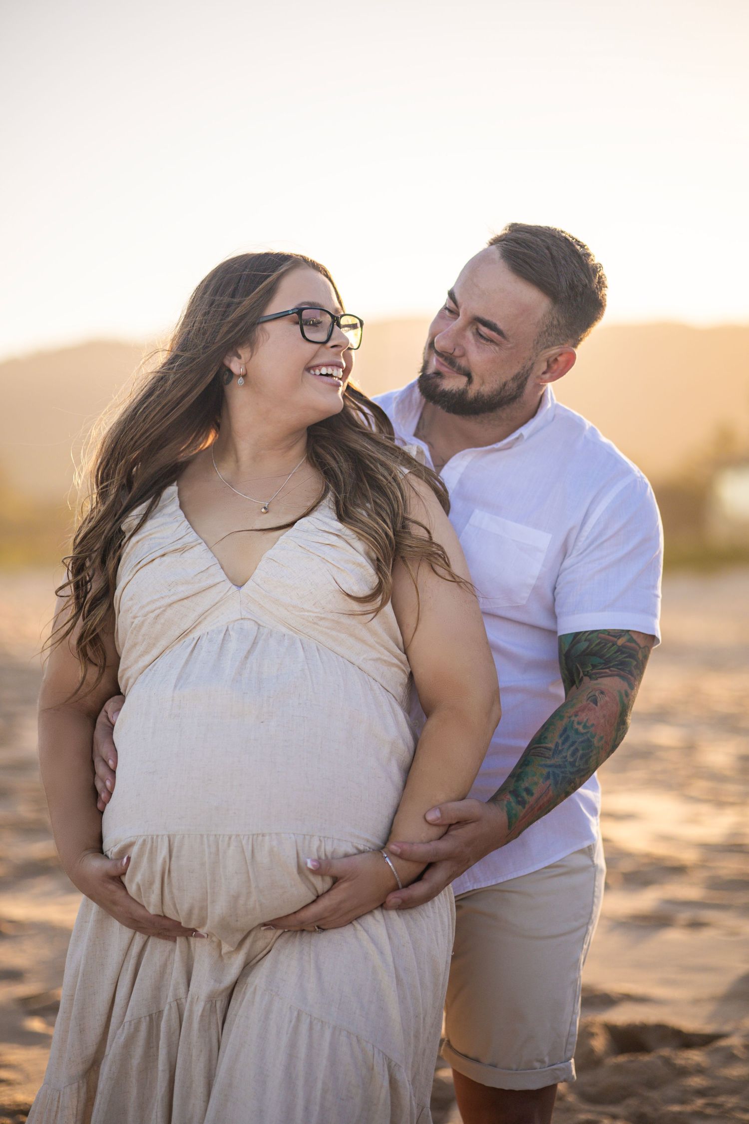Happy pregnant couple embracing on sandy beach during golden sunset.