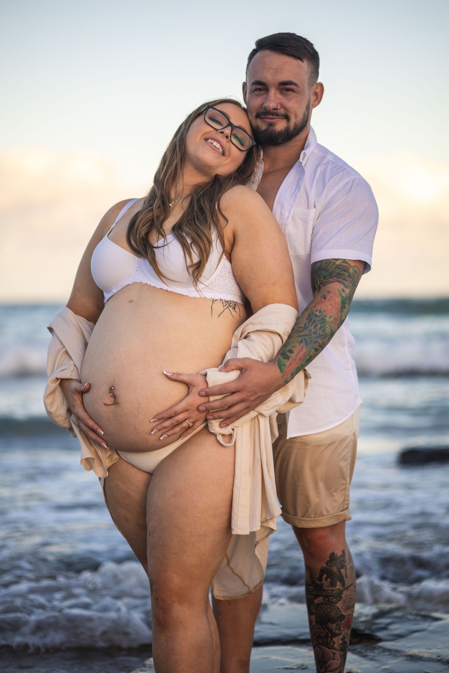 Couple embracing on the beach at sunset, man holding pregnant partners baby bump tenderly.
