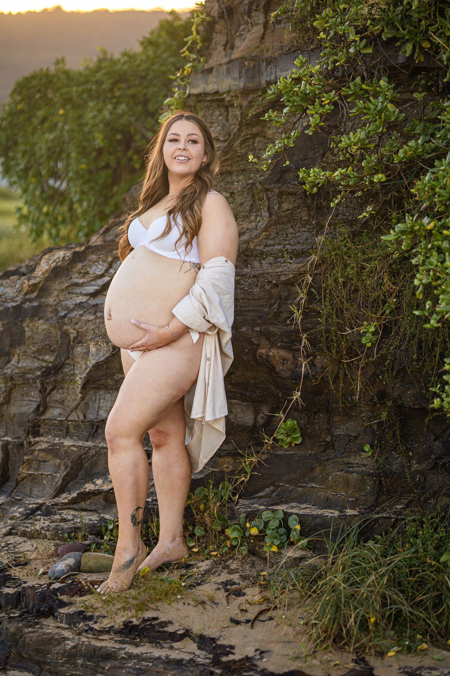 Pregnant woman in white bra and open shirt posing by rocky cliff with greenery.