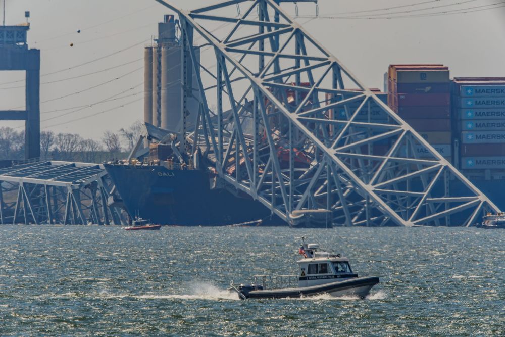 A small patrol boat cruises past towering electrical transmission towers and cargo containers along an industrial waterfront.