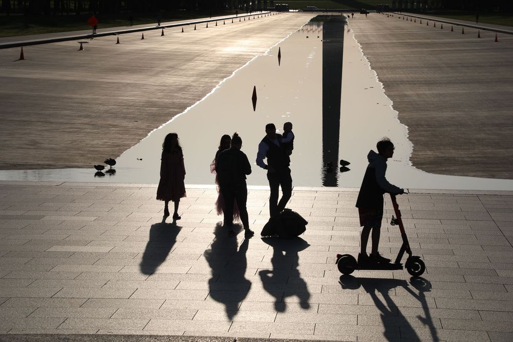 Long shadows cast on a concrete plaza as silhouetted people walk and ride scooters in the evening light.