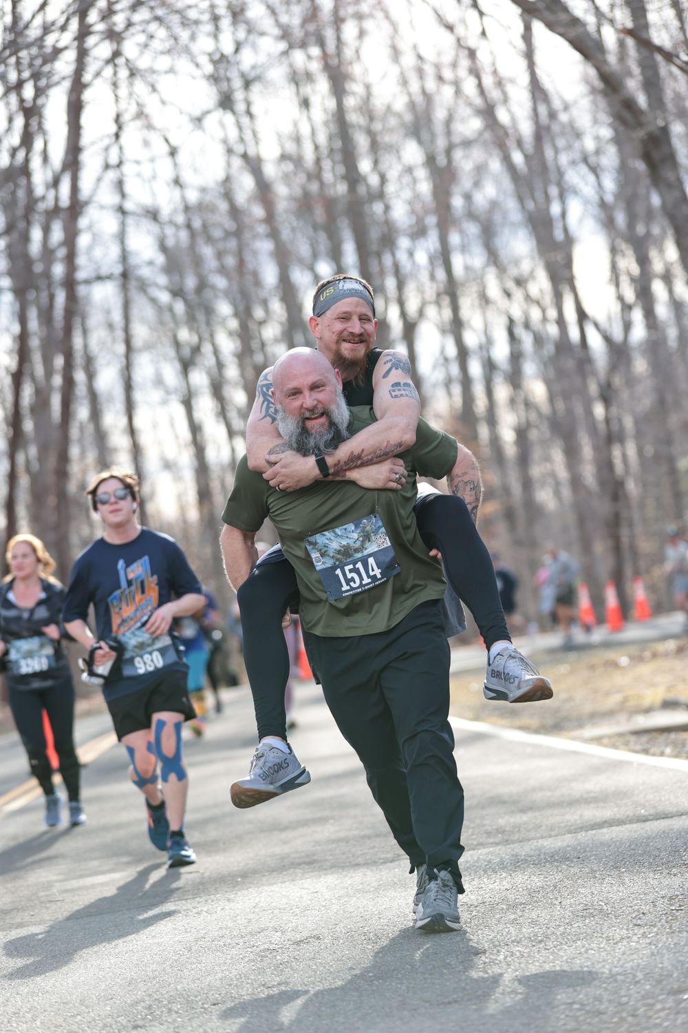 Two runners share a playful moment during a race as one gives the other a piggyback ride on a wooded path.
