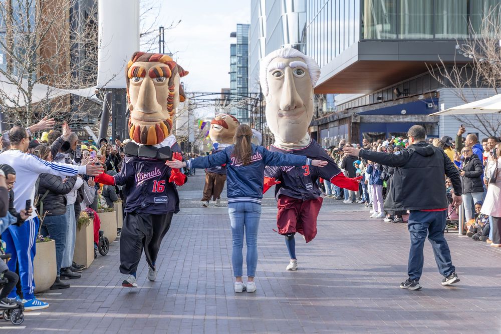 Performers wearing oversized heads entertain crowds on an urban sidewalk during a street festival.