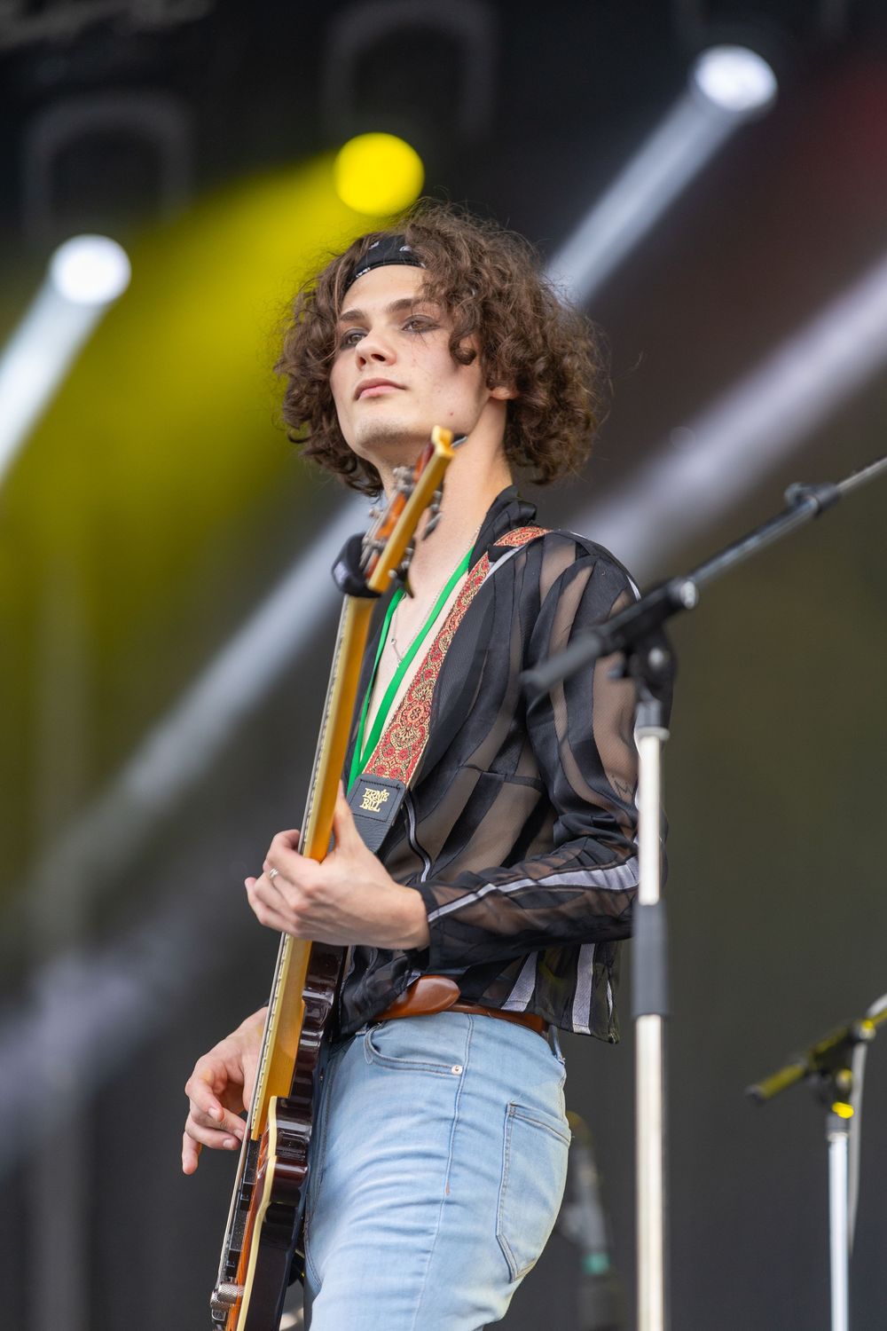 A musician performs on stage with a guitar wearing a sheer black shirt and jeans under dramatic concert lighting.