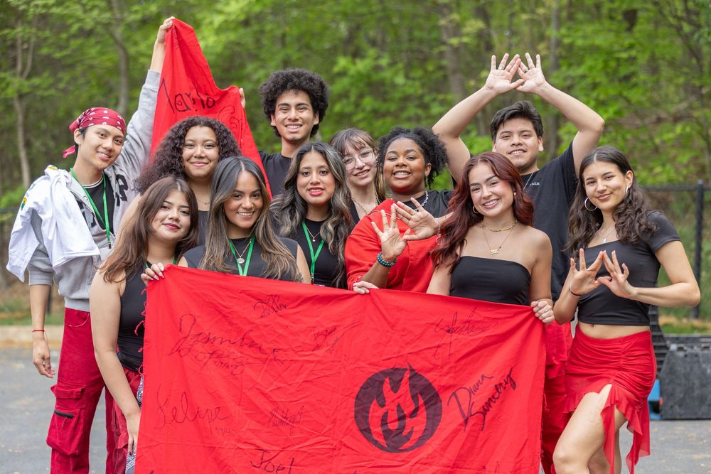 A group poses outdoors holding a red flag with enthusiasm while dressed in red and black attire.