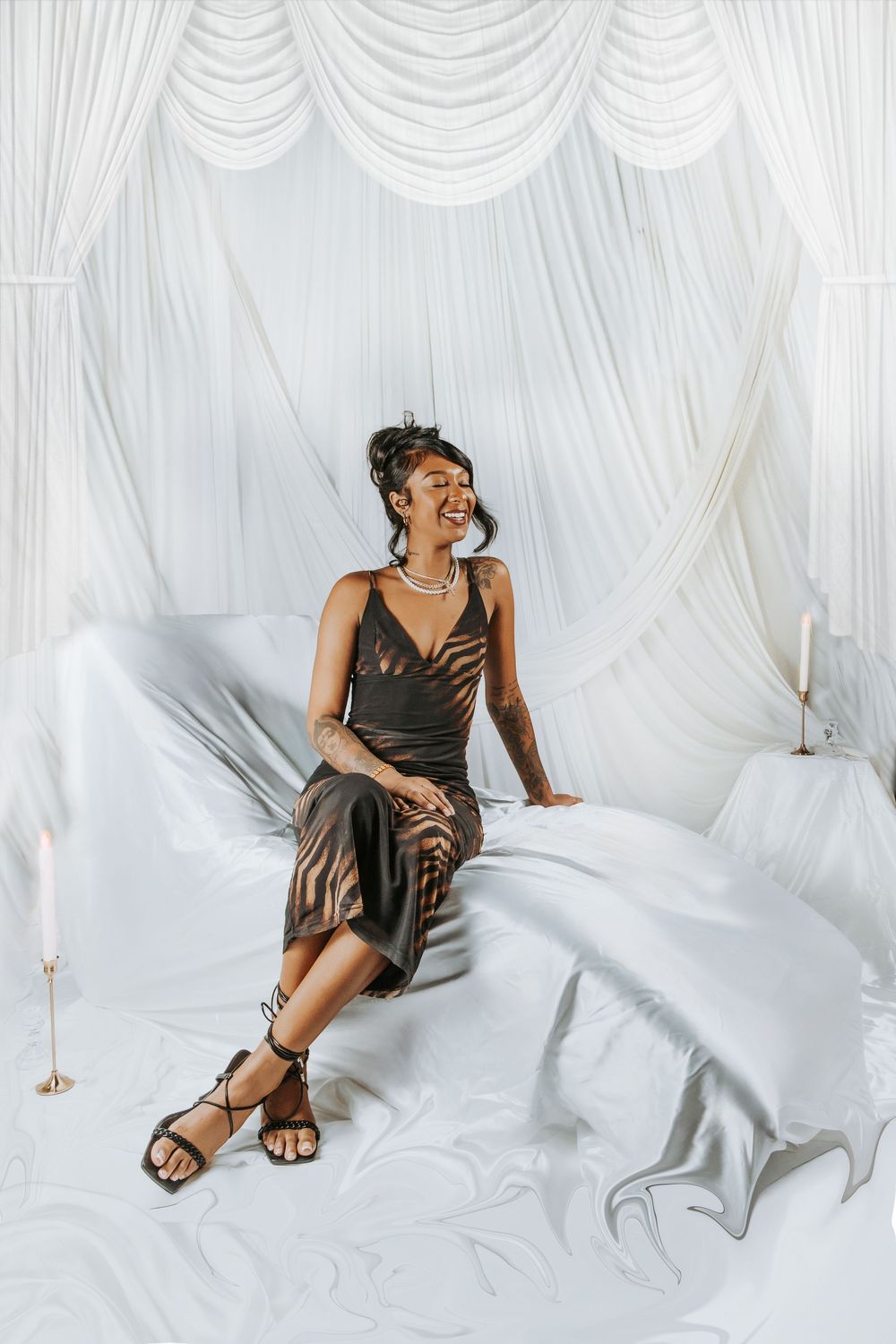 A woman in a bronze satin dress and strappy sandals sits laughing on a white draped bed in an elegant studio setting.