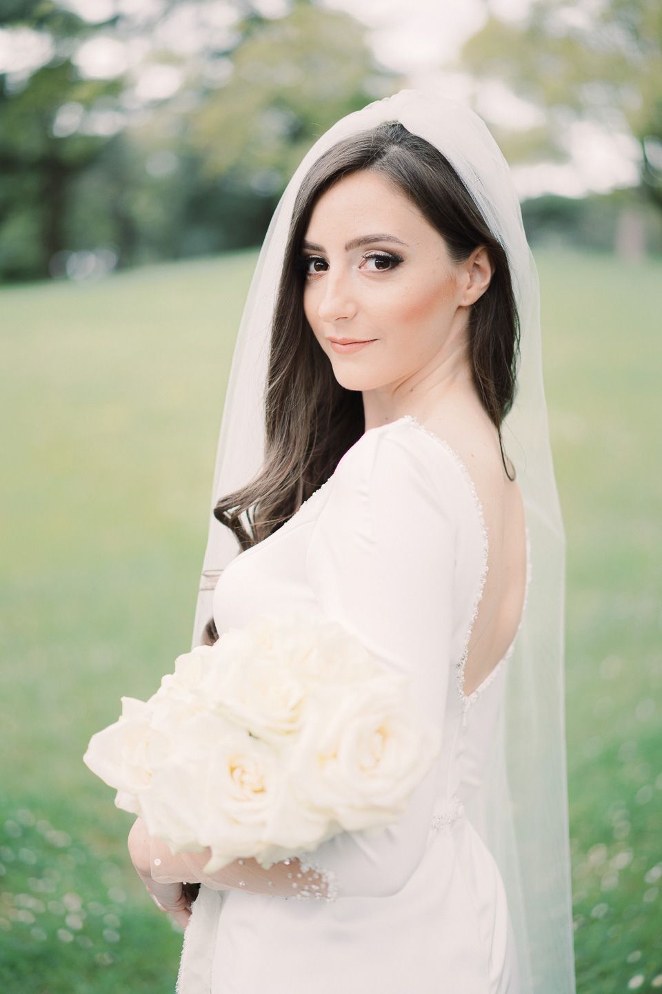 Bride smiling at the camera confidently, holding her bouquet of white roses, and wearing a wedding dress with a deep back neckline