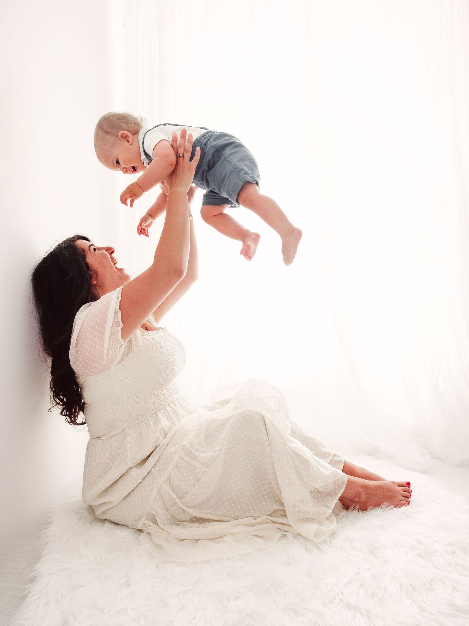 A mum in a cream dress sits and holds her one year old baby boy up in the air, they are both laughing.