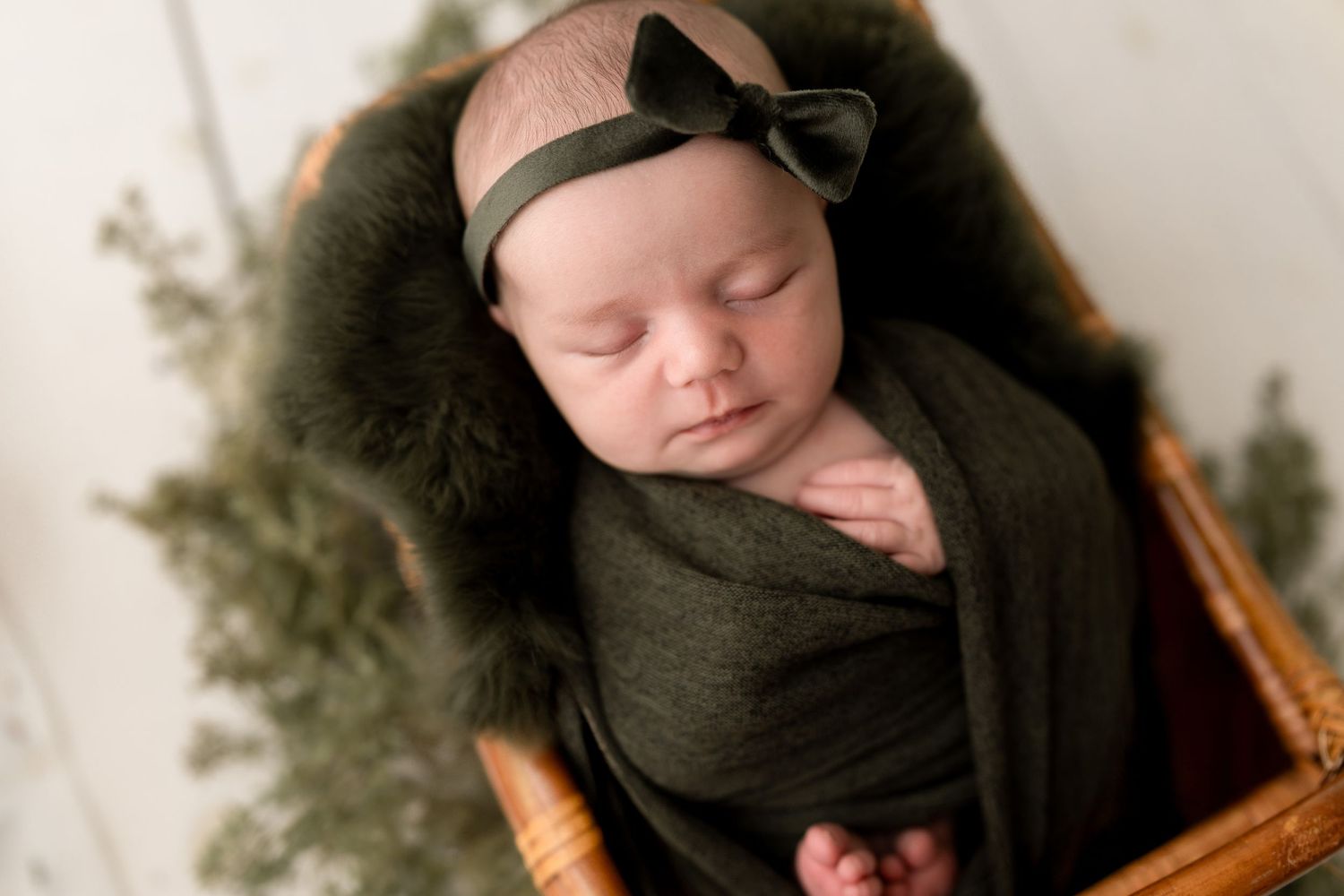 Sleeping newborn wrapped in olive green swaddle with bow headband in wooden basket with greenery.