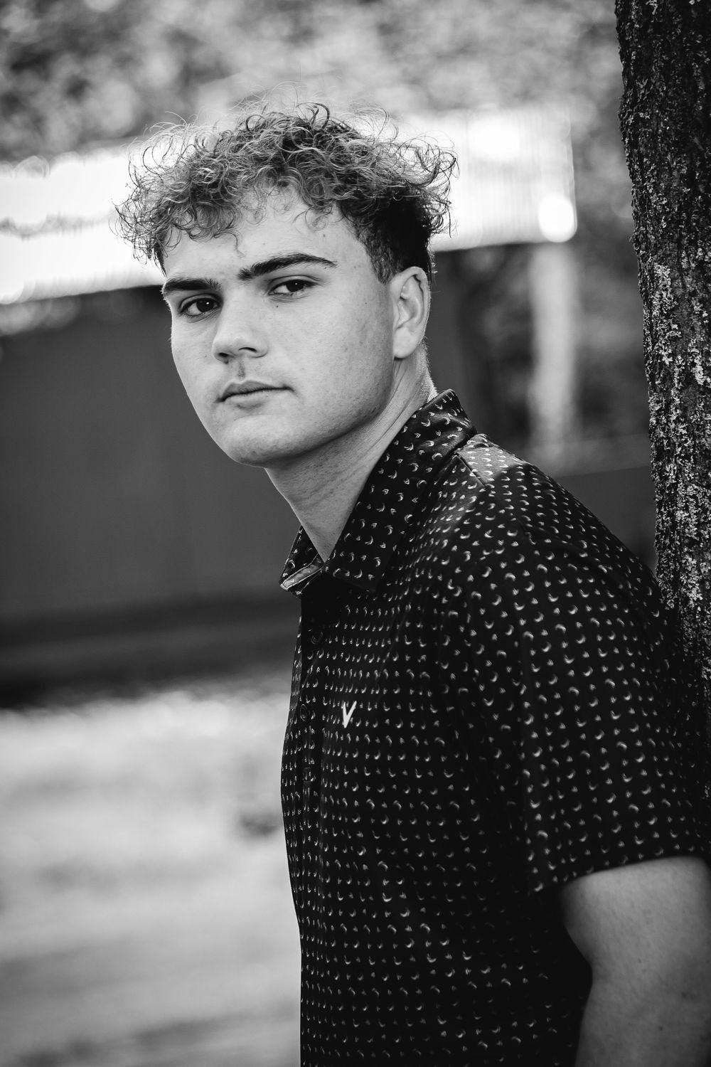 Black and white portrait of a young man with curly hair wearing a patterned shirt, leaning against a tree outdoors.