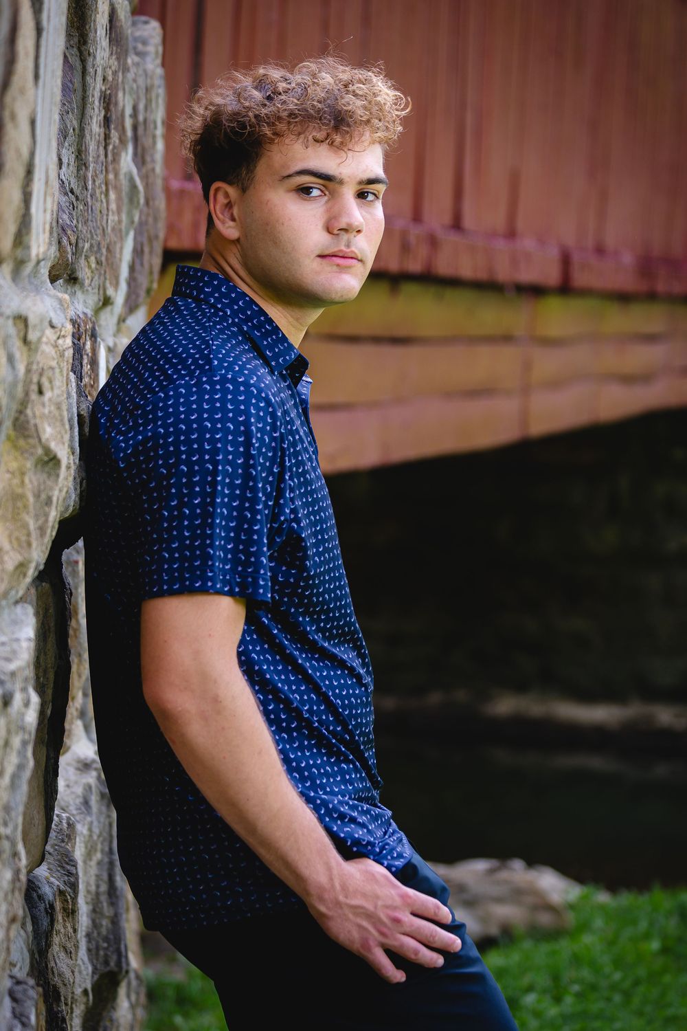 Young man with curly hair leans against stone wall in blue patterned shirt, posing outdoors near a red covered bridge.