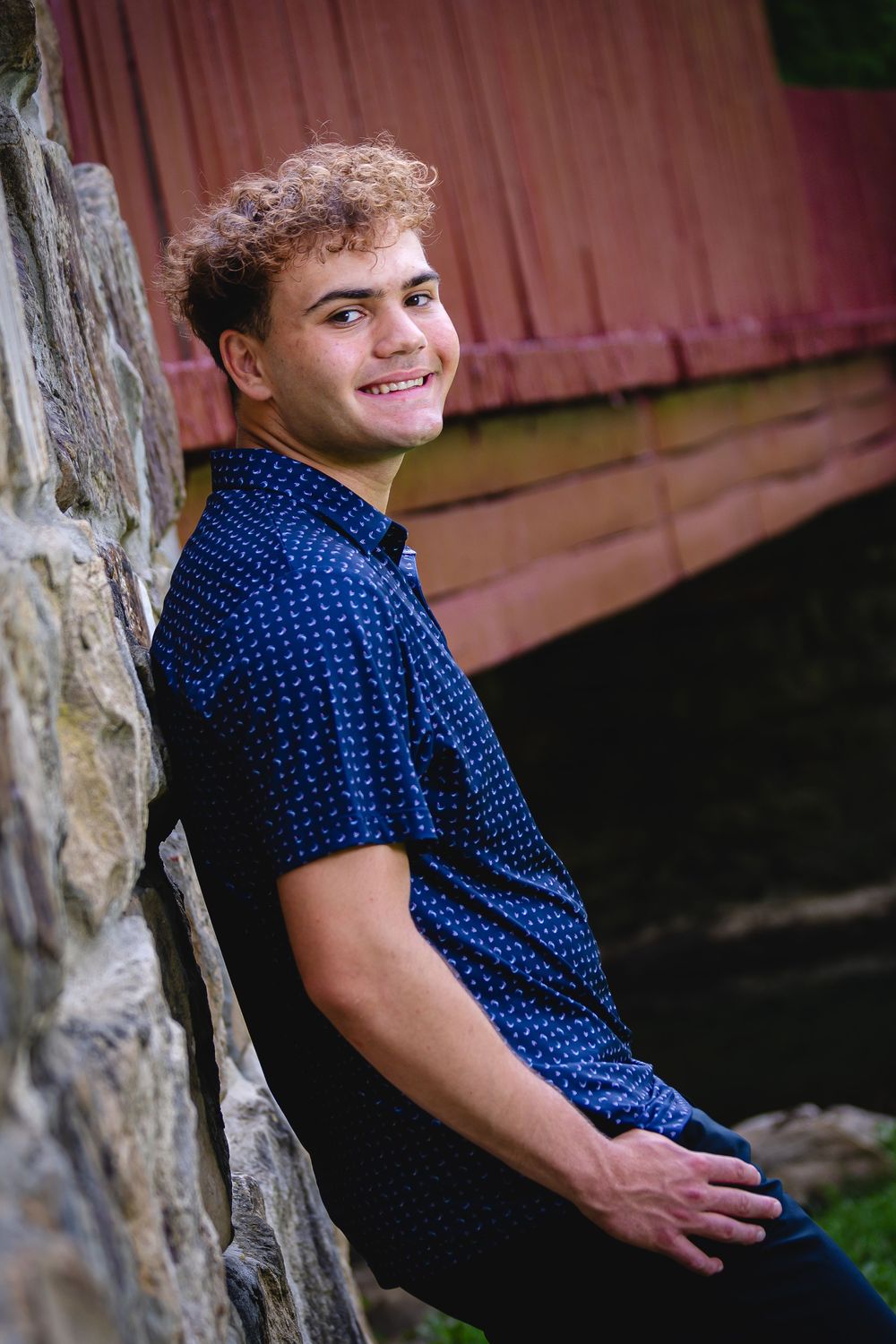 Smiling young man with curly hair in a blue shirt leaning against a stone wall near a red covered bridge.