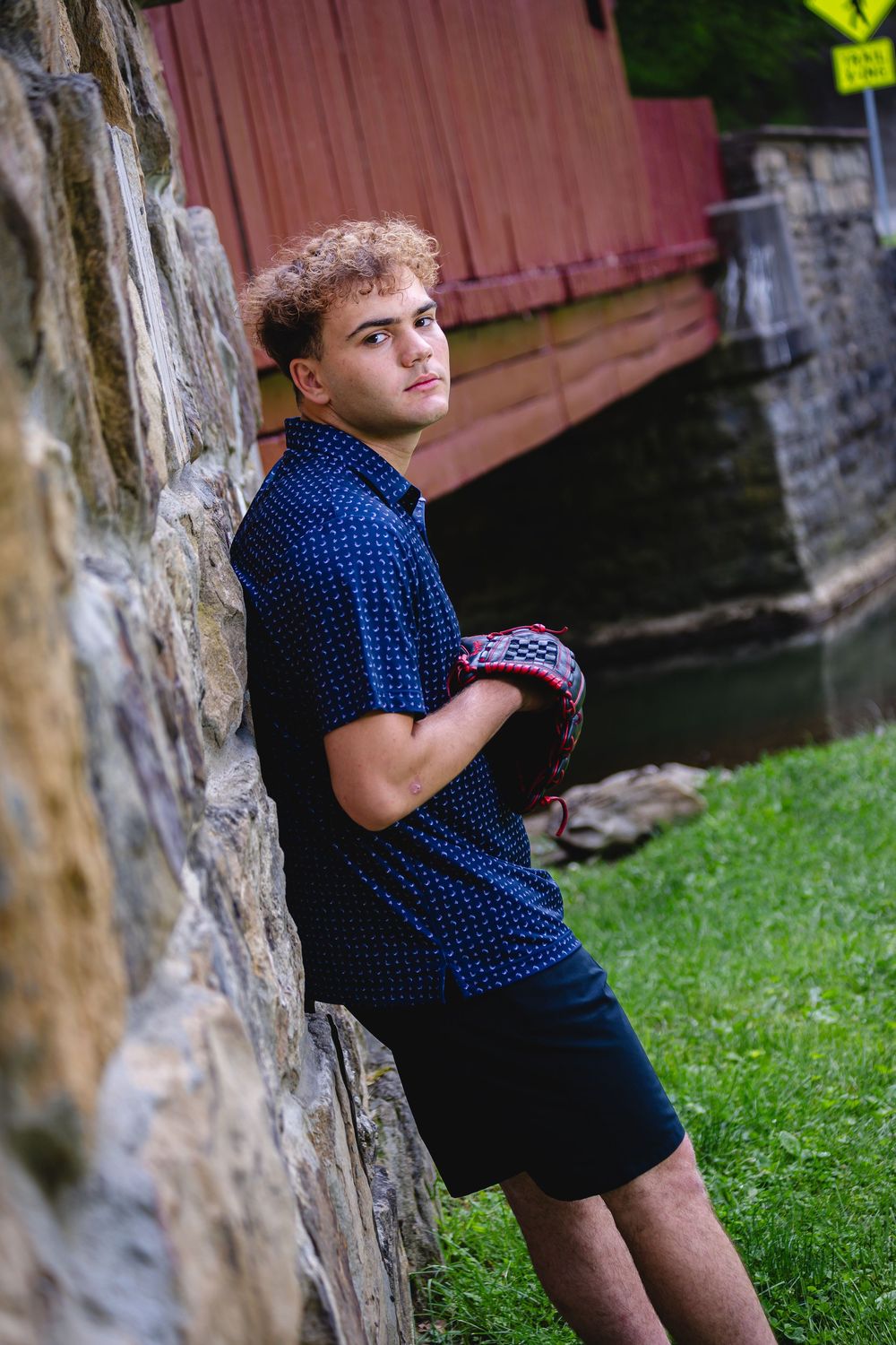 Young man in navy blue shirt and shorts leaning against stone wall near a red covered bridge.
