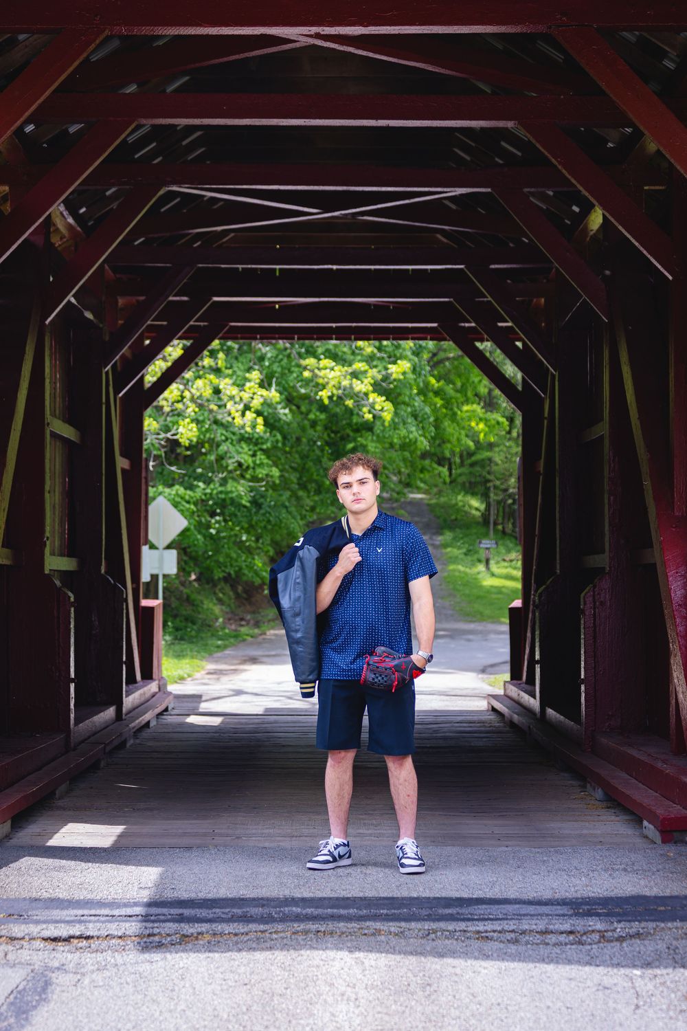 Young man in navy blue outfit standing inside a red covered wooden bridge with lush green trees visible beyond.