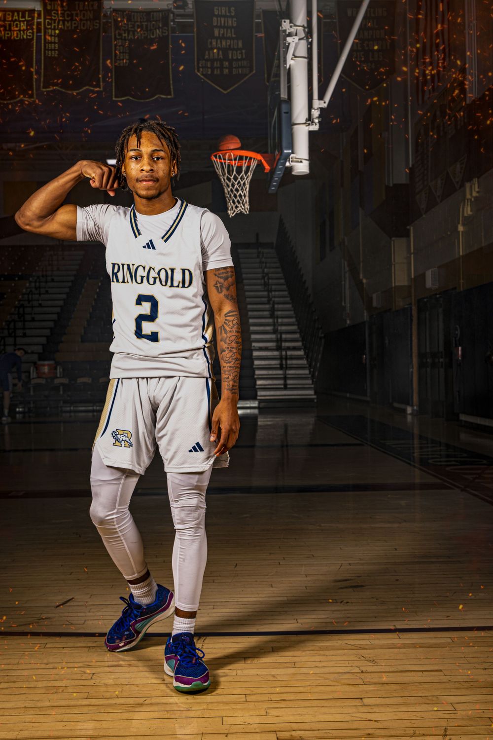 Basketball player wearing a white Kinggold #2 jersey poses on wooden court with a basketball hoop in background.