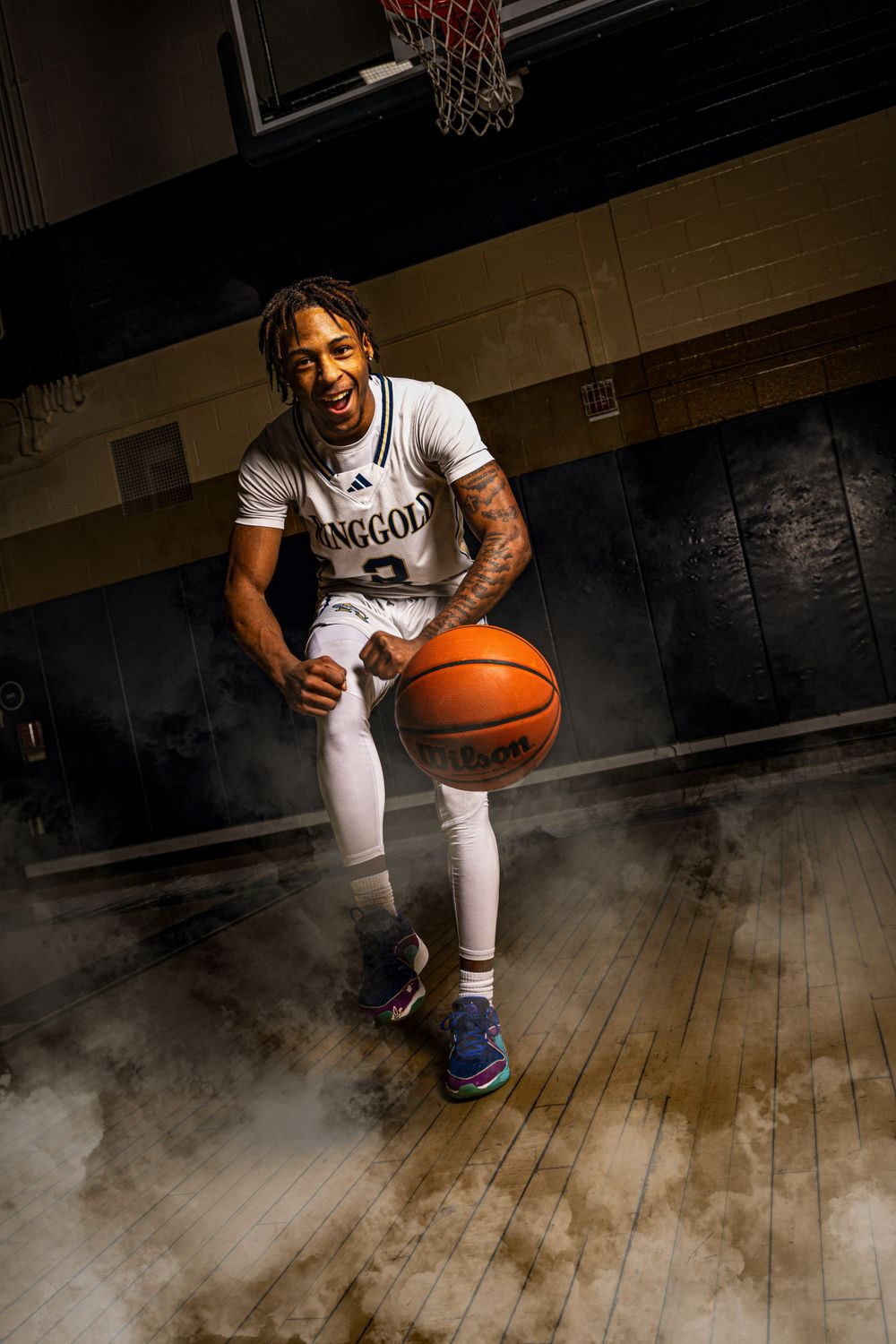 Basketball player in white uniform dribbles ball on wooden court with dramatic fog effect and basketball hoop above.