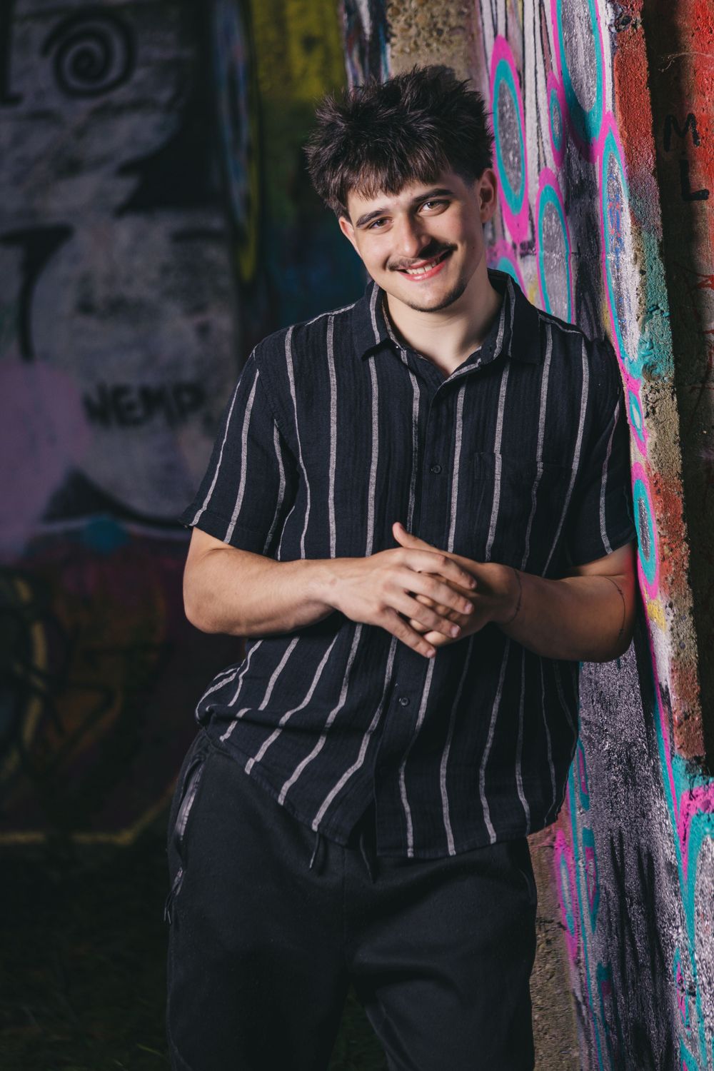 Young man with mustache smiling in striped black shirt leaning against colorful graffiti wall.
