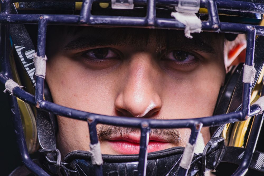 Close-up of a young male football player wearing a navy blue helmet and face guard, intense focused expression.