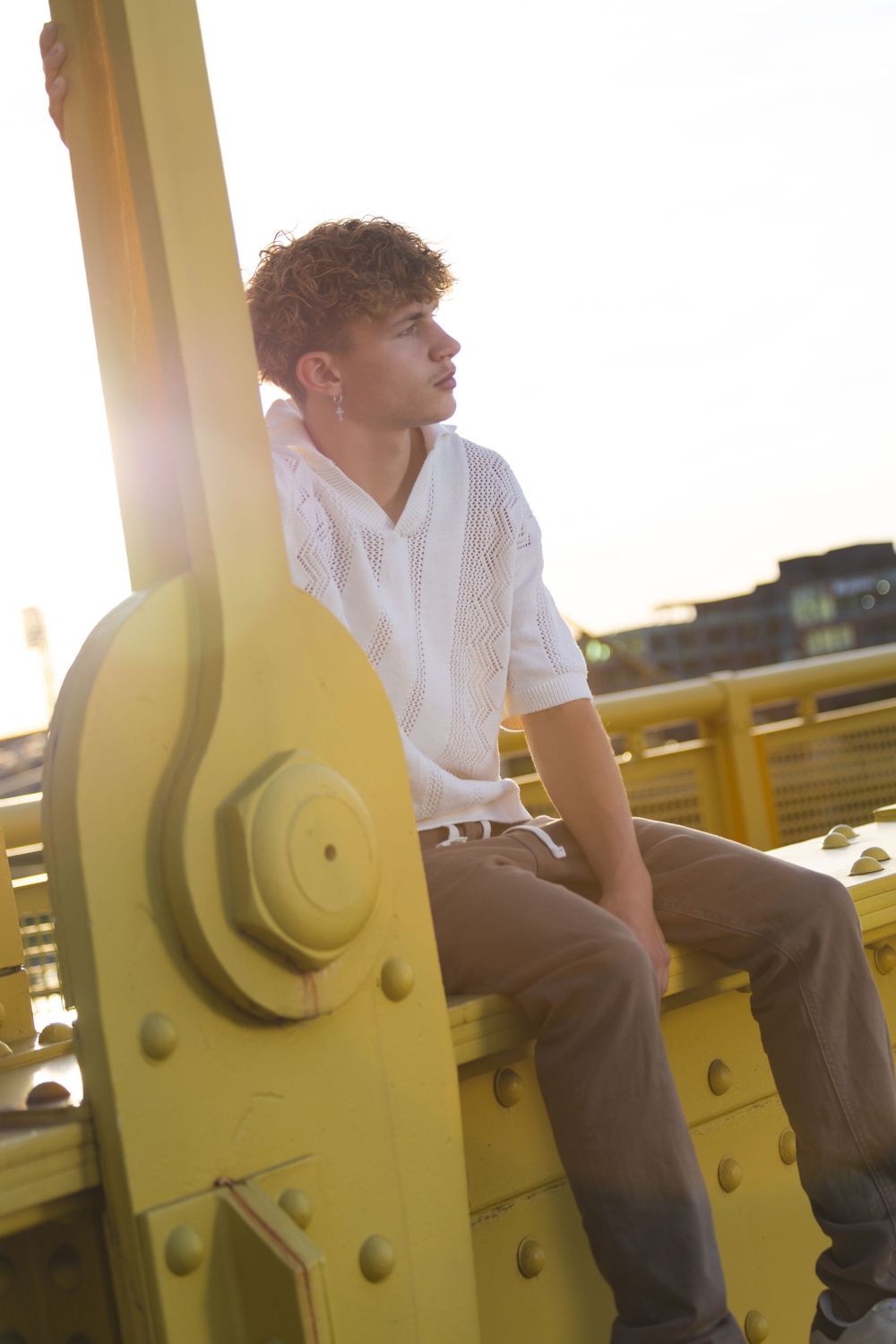 Young man in white shirt and khaki pants sitting on yellow steel bridge structure at golden hour.