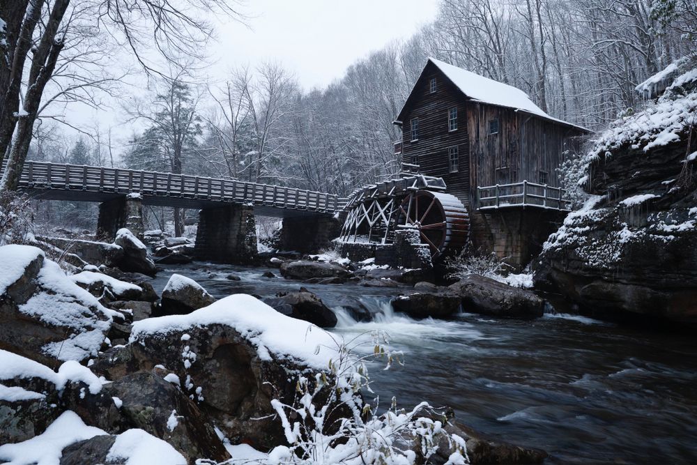 Snow covered grist mill and wooden bridge over rushing water in winter landscape.