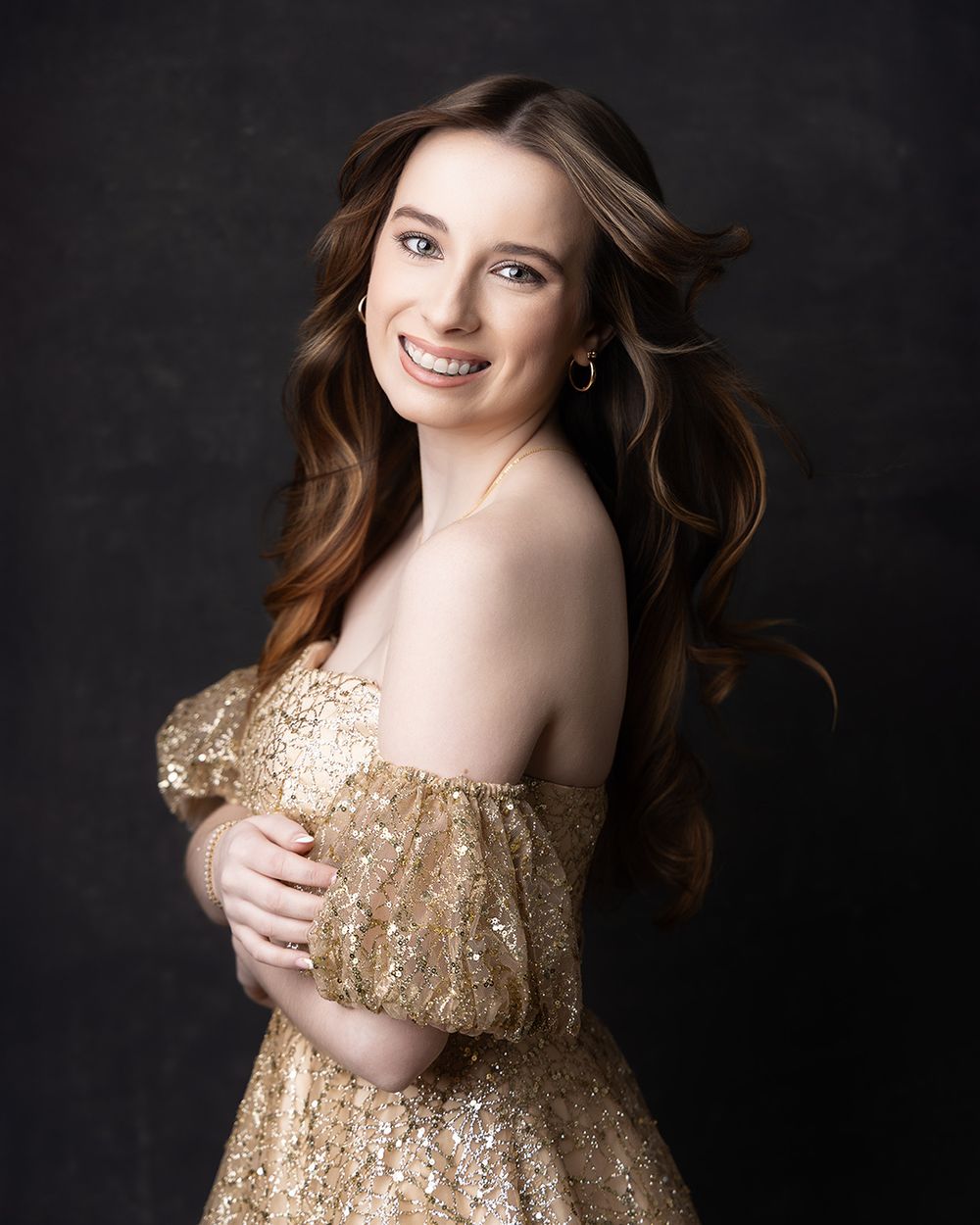 Young woman smiling in a gold sequin off-shoulder gown with wavy auburn hair against a dark background.
