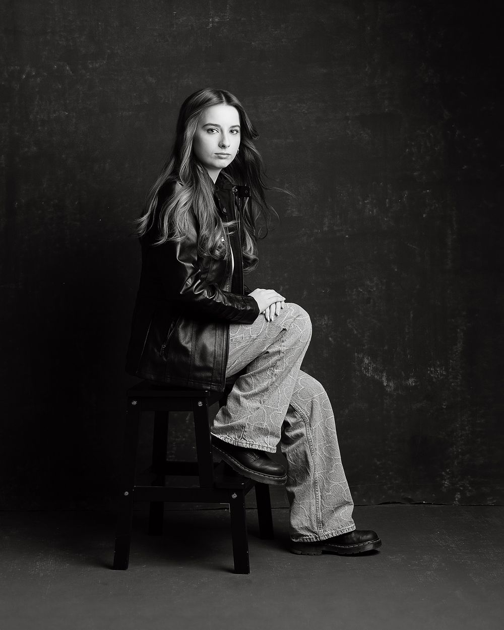Black and white studio portrait of a young woman sitting on a wooden stool, wearing jeans and a dark jacket.