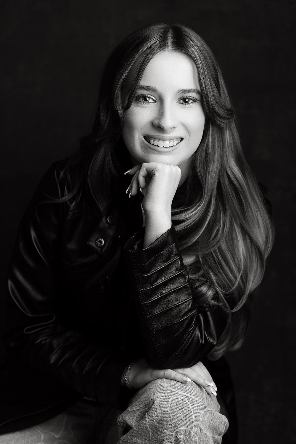 Black and white portrait of a smiling woman with long hair in a leather jacket, resting chin on hand.