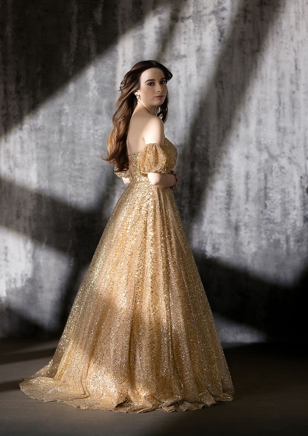 Woman in a stunning gold sequin off-shoulder ball gown posing elegantly against a textured gray wall.