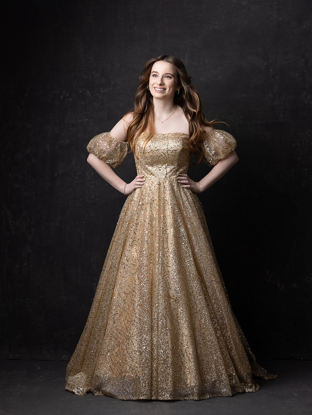 Young woman in a gold sequin ball gown with puff sleeves, posing confidently against a dark background.