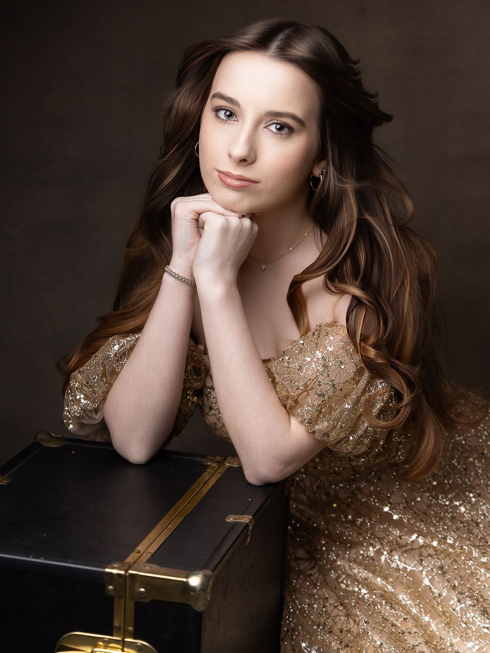 Young woman in gold sequin dress with long wavy brown hair, posing elegantly with hands clasped near chin.