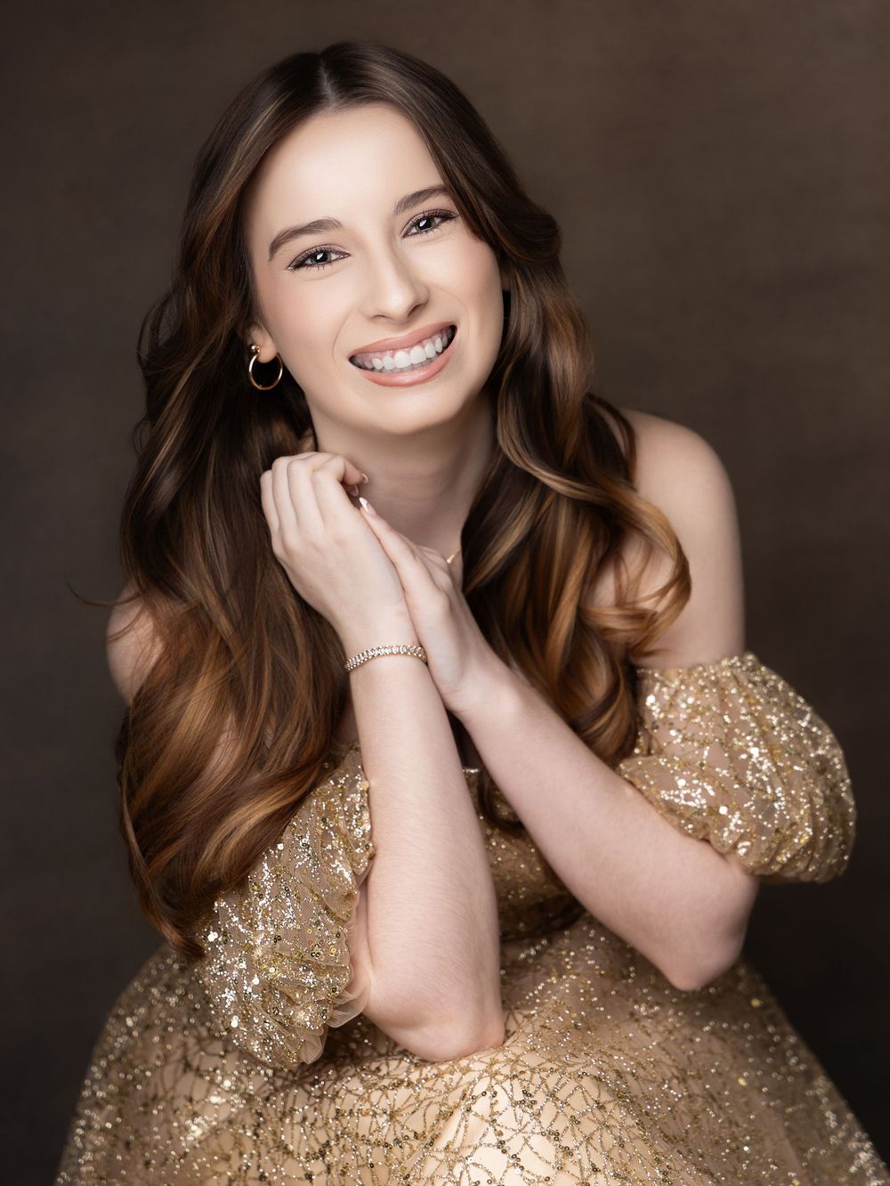 Young woman in gold sequin off-shoulder dress smiling warmly, with long brown hair and gold jewelry.