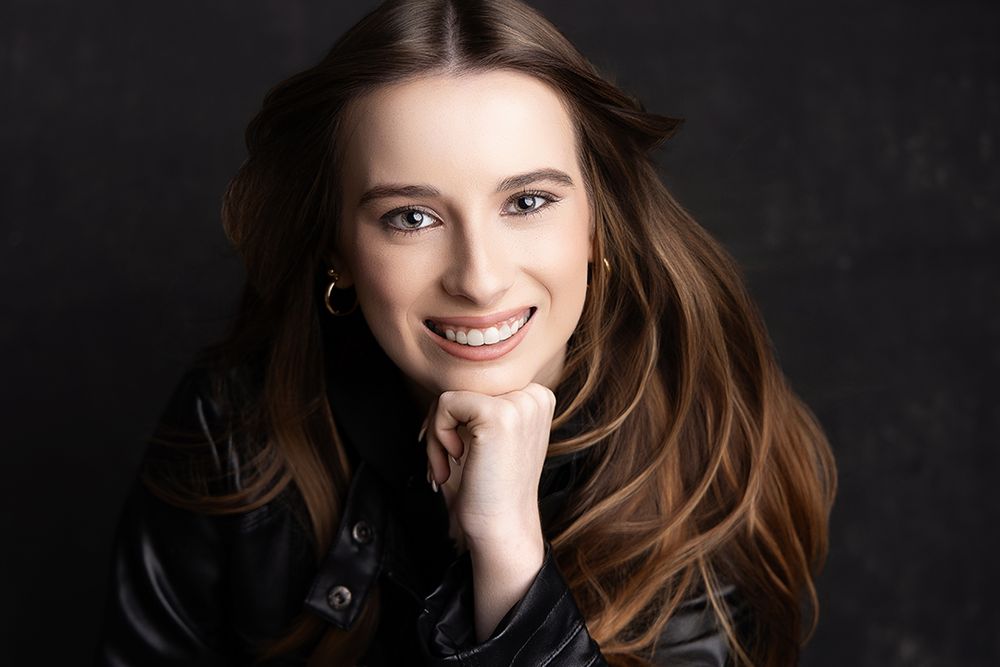 Smiling young woman with long brown hair wearing a black leather jacket, posing confidently against a dark background.