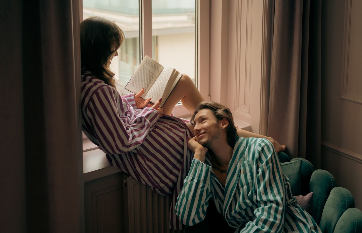 Friends in matching striped loungewear share a laugh while reading by a sunlit window.