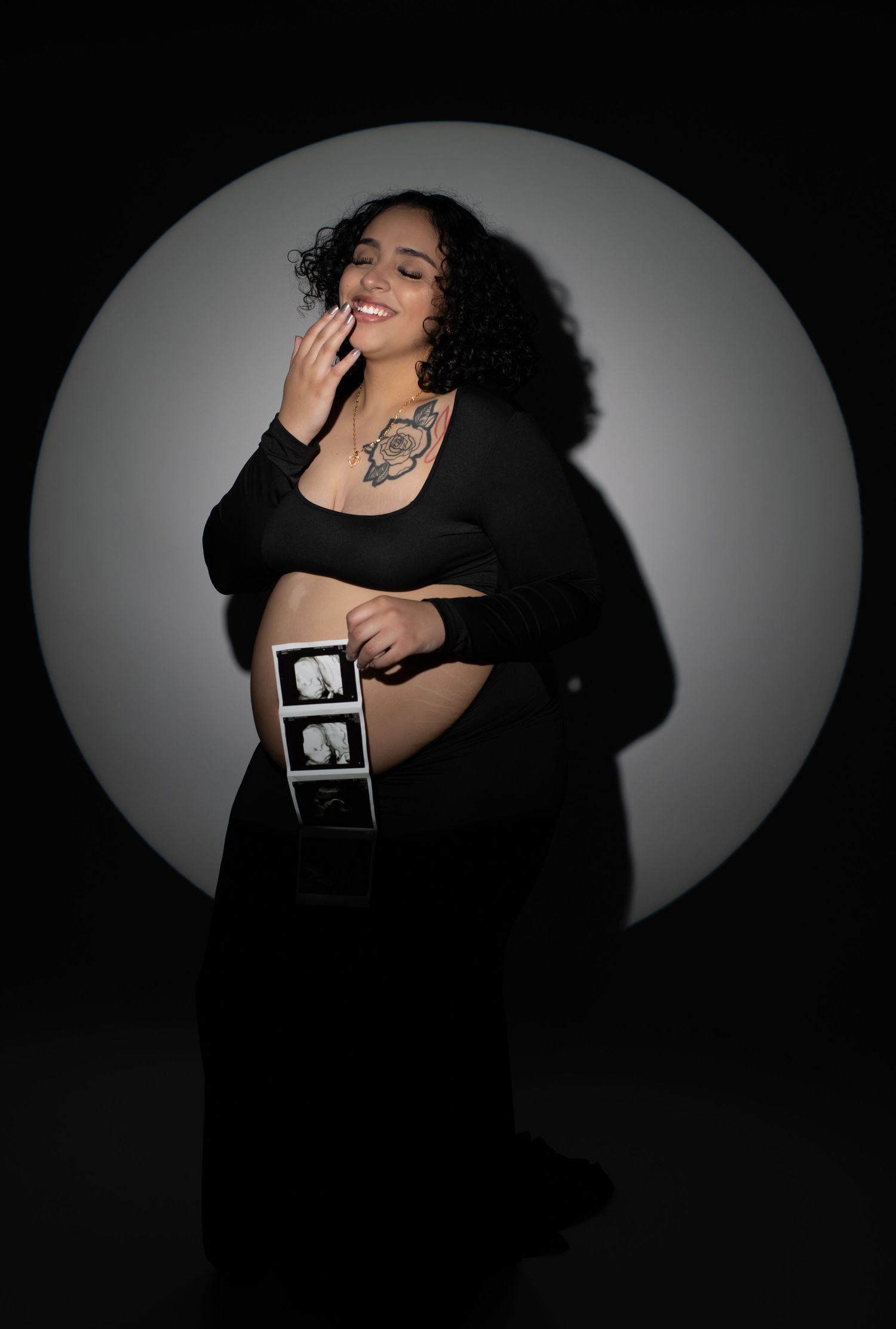 Smiling pregnant woman in black outfit holds ultrasound photos against belly in dramatic circular light studio.