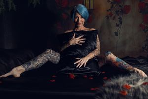 A 50+ woman with elaborate leg tattoos poses on a dark backdrop with scattered red petals in this romantic boudoir photo taken in Fort Worth