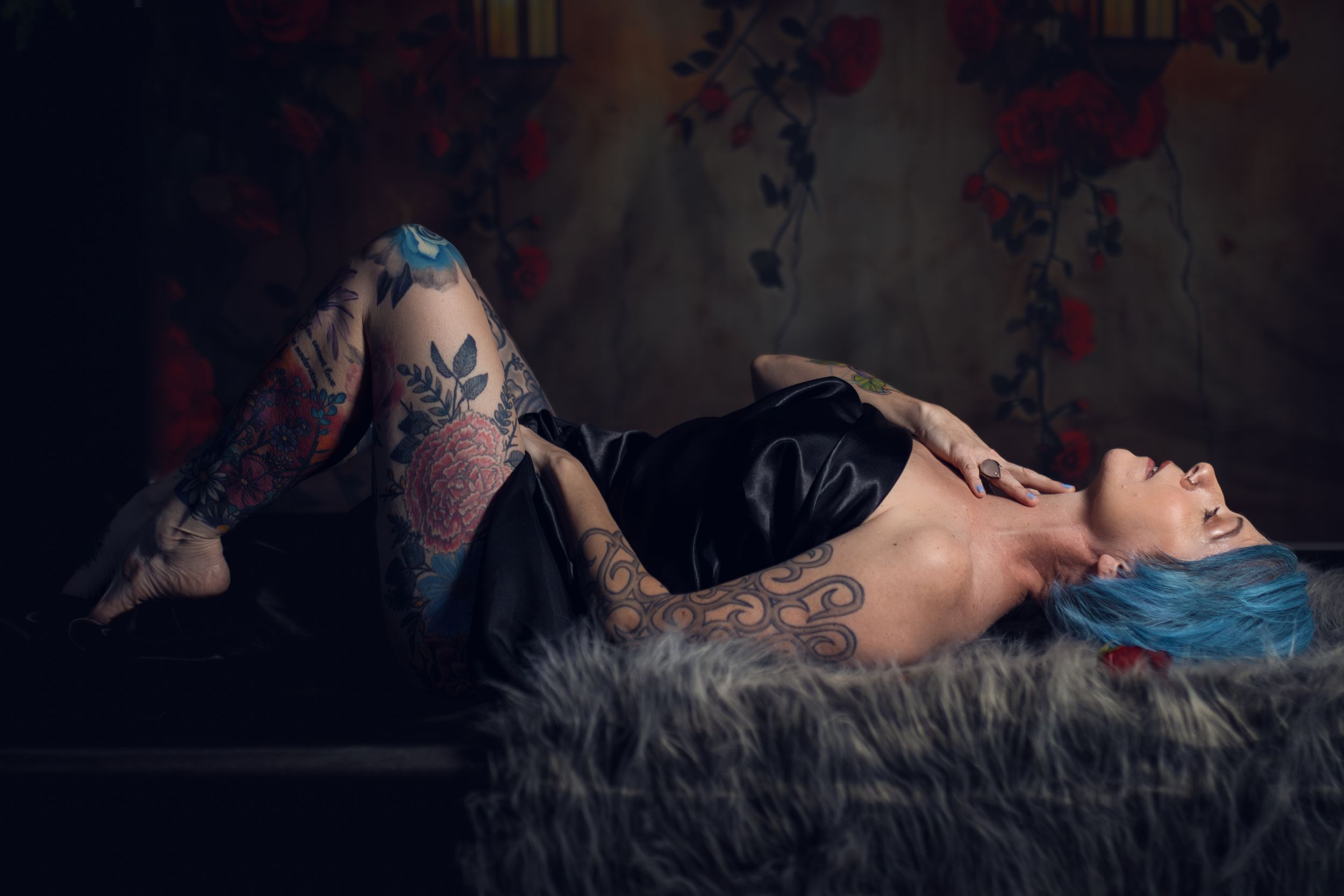 A person with blue hair and tattoos reclines on a dark couch wearing a black dress against a moody backdrop in this Fort Worth boudoir photo