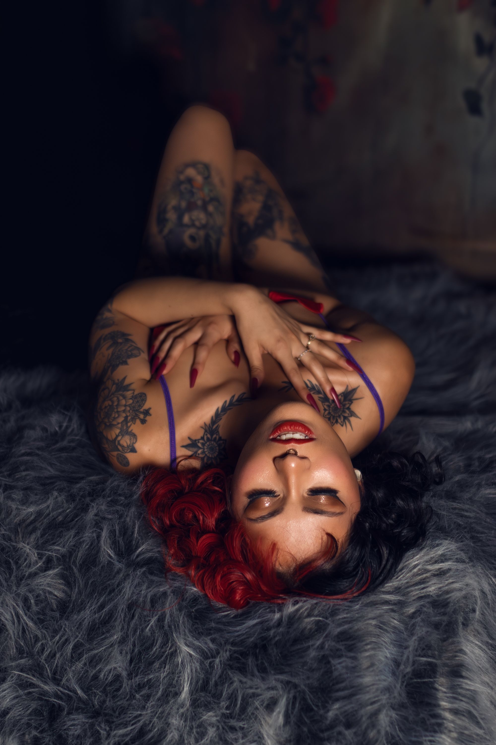 Person with vibrant red hair and tattoos poses on gray fur in artistic boudoir photo shoot in Keller, Texas.