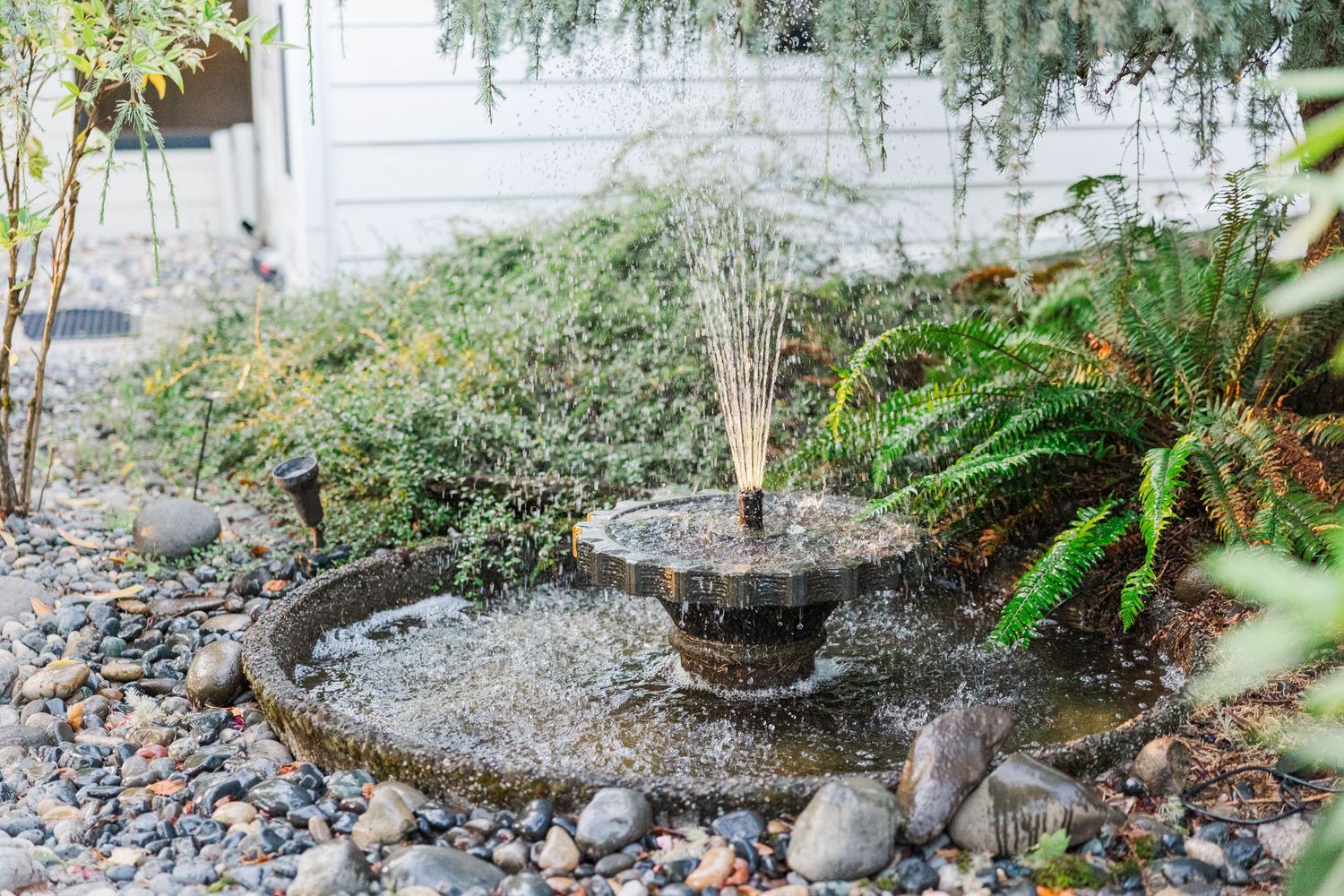 Decorative stone water fountain surrounded by rocks, ferns, and greenery in a peaceful garden.