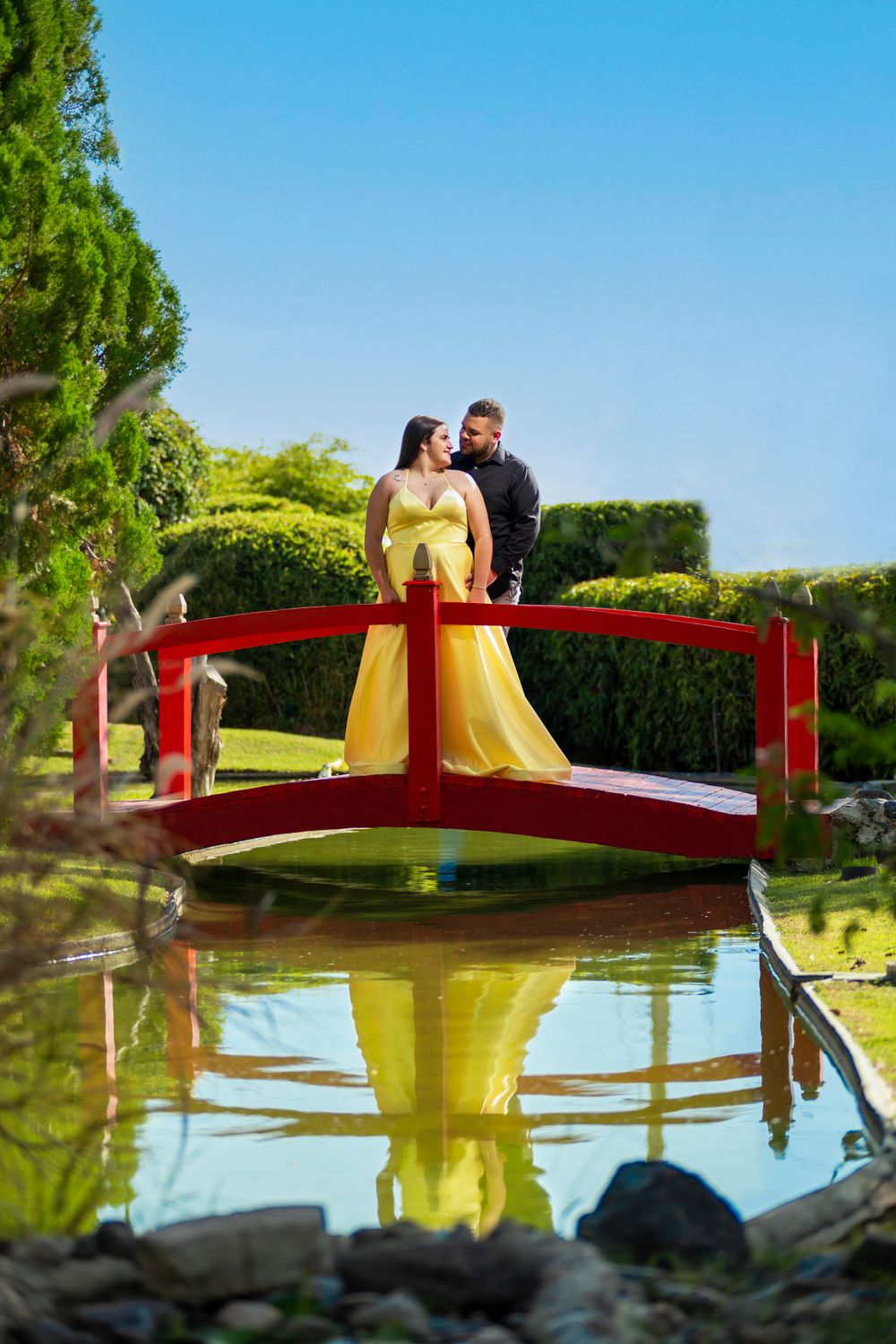 A couple in formal attire poses on a red bridge over a reflective pond in a manicured garden.