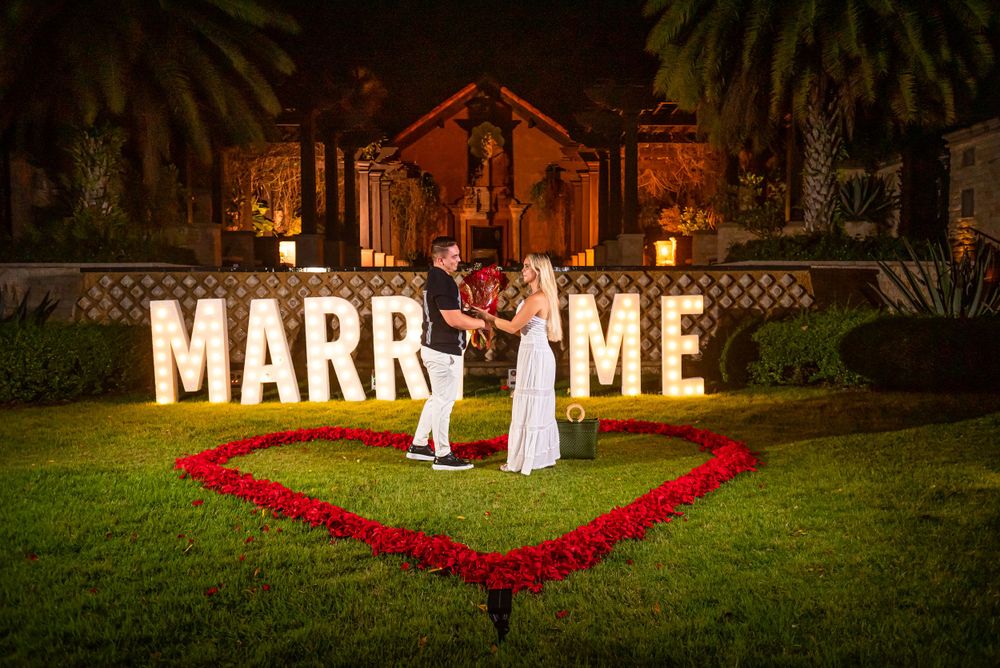 Illuminated 'MARRY ME' proposal setup with red rose heart shape and palm trees at night.