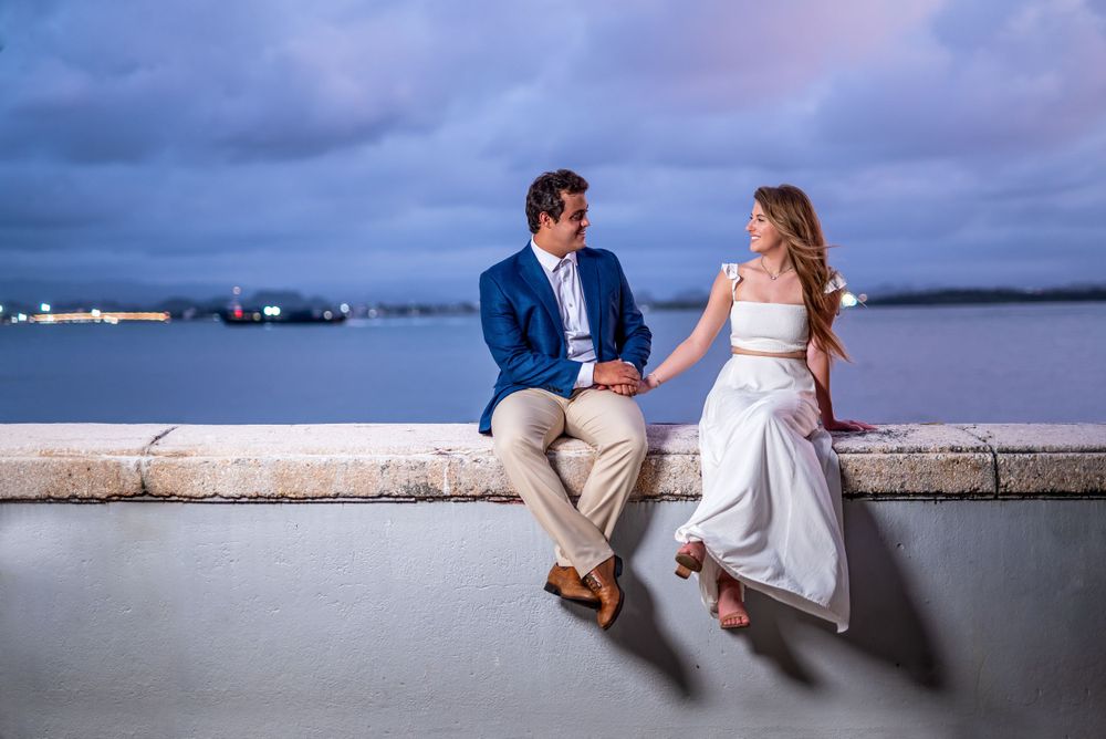 A couple sits casually on a waterfront wall at dusk with harbor views in the background.