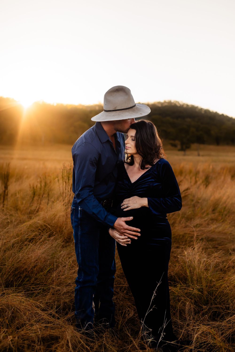 Couple embraces in golden field at sunset while wearing navy blue and black clothing.