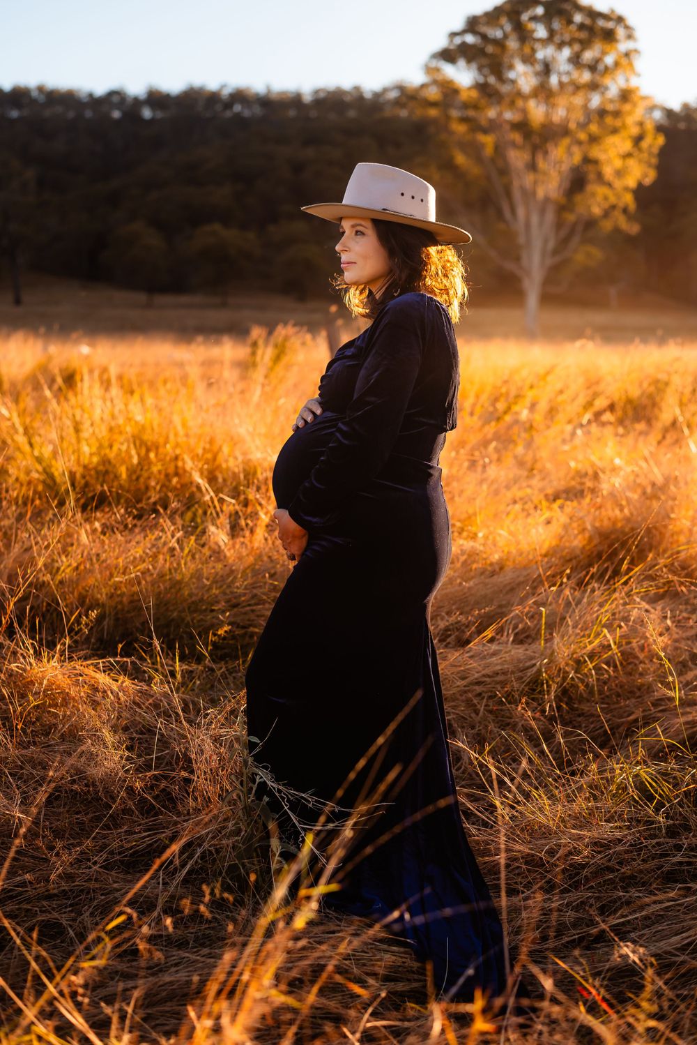 Woman in black dress and hat stands in glowing field during sunset.