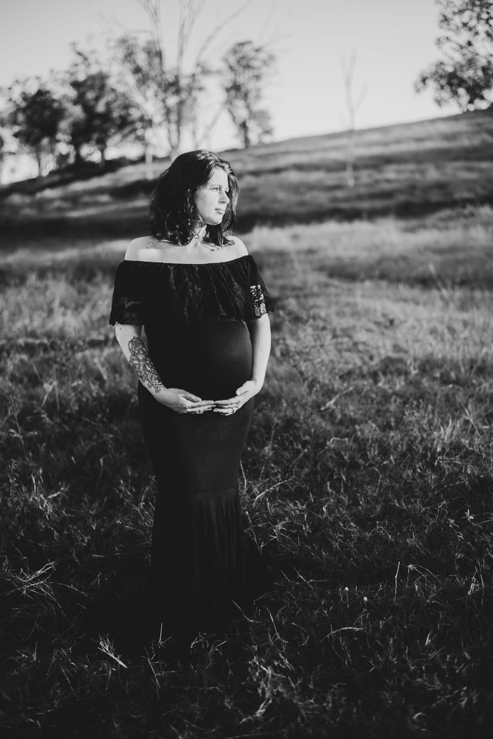 Woman in black off-shoulder dress poses in grassy field for maternity photo.