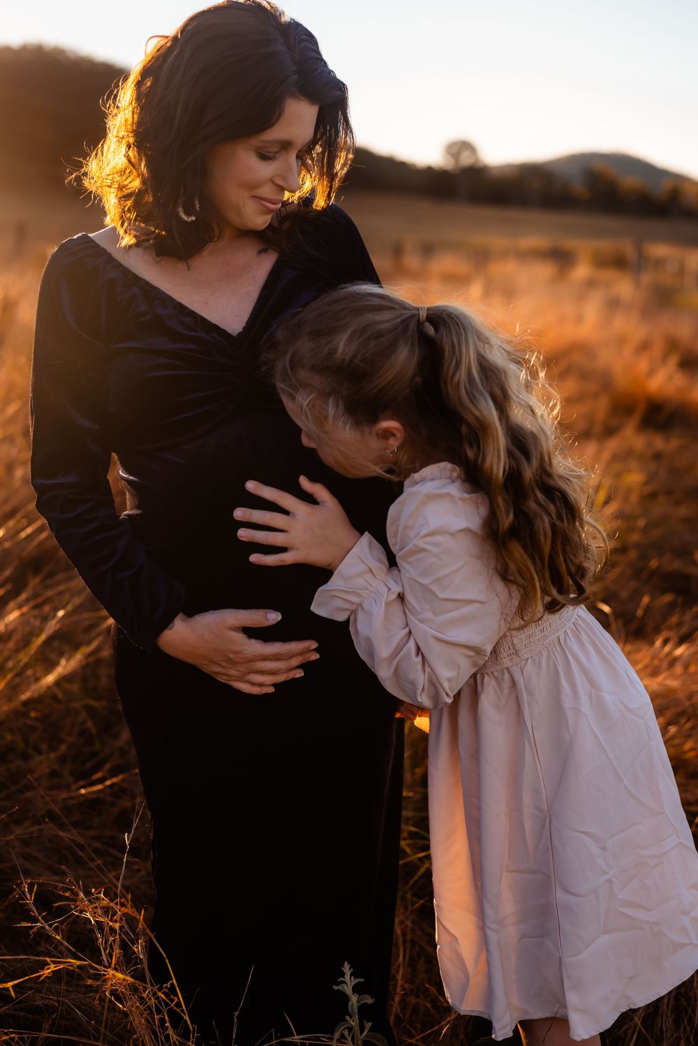Child hugs mother's baby bump during sunset maternity photoshoot in field.