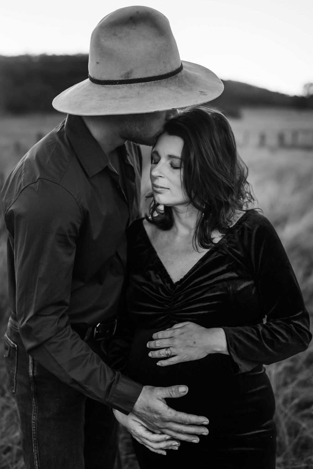 Intimate maternity portrait of couple in black clothing against rustic field backdrop.