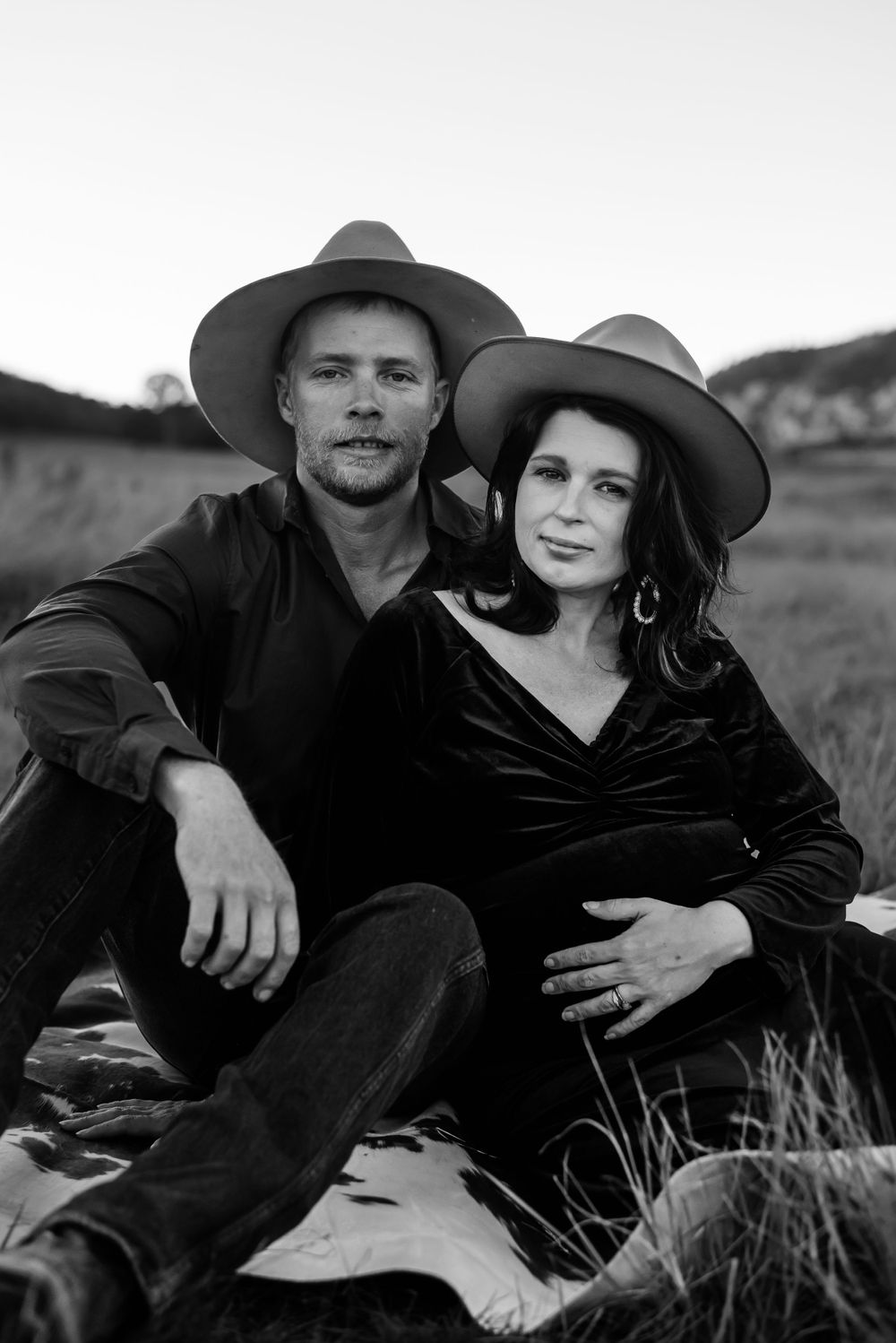A classic black and white portrait of a couple in western hats sitting together in a grassy field.