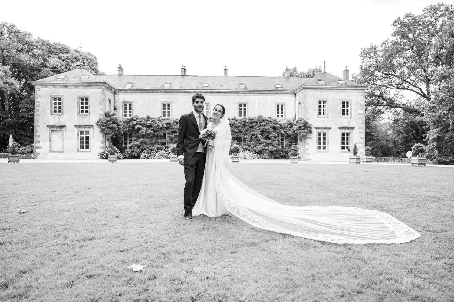 Black and white wedding photo of a couple standing in front of an elegant manor house.