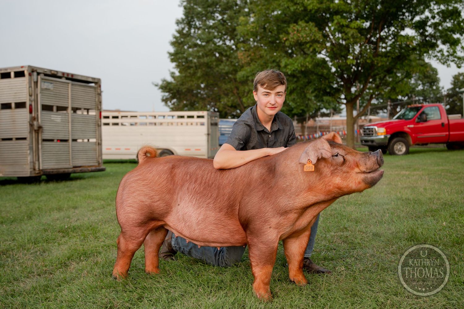 An exhibitor poses with their red show pig on grass with livestock trailers in the background.